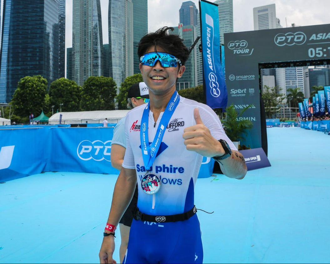 A male athlete wearing blue and white cycling apparel, blue sunglasses, and a medal stands at the finish line of the PTO Asian Open cycling event, with the Singapore city skyline in the background.