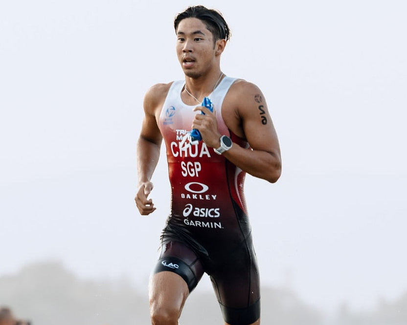 A male athlete running barefoot on a sandy beach wearing a red, white, and black athletic triathlon suit with visible ASICS branding.