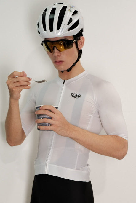 Person wearing a white cycling jersey and black shorts with a helmet and sunglasses, holding a coffee cup.
