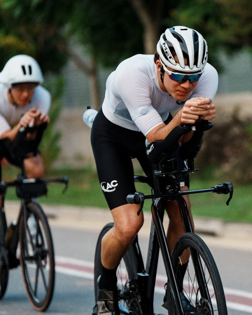 Three cyclists in a time trial group riding on a road, wearing white jerseys, black shorts, and aerodynamic helmets and sunglasses.