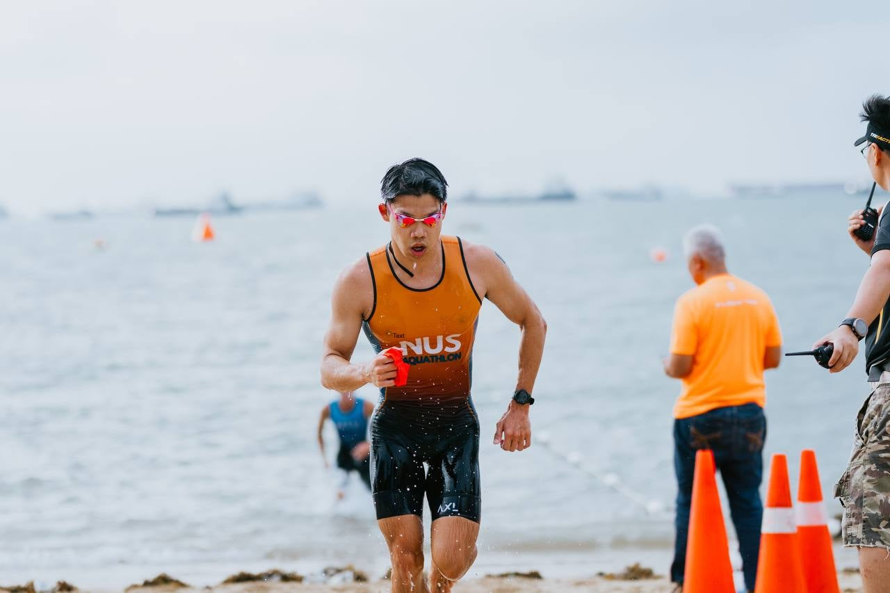 A man running on a beach during an athletic event, wearing a black and orange athletic tank top with 'NUS' printed on the front, black running shorts, and red-tinted sunglasses. Other people and orange cones are visible in the background near the water.